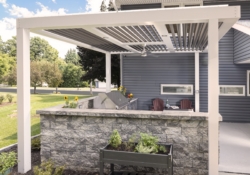 Pergola-covered Jakab outdoor kitchen with stone surround, grill, and seating by Mother Hubbard custom cabinetry.