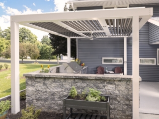 Pergola-covered Jakab outdoor kitchen with stone surround, grill, and seating by Mother Hubbard custom cabinetry.